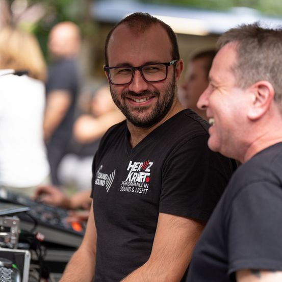 Zwei Männer lächeln und unterhalten sich bei einer Outdoor-Veranstaltung, einer trägt ein schwarzes T-Shirt mit dem Text 'HEIZ KRAFT' und der andere ein schwarzes Shirt.
