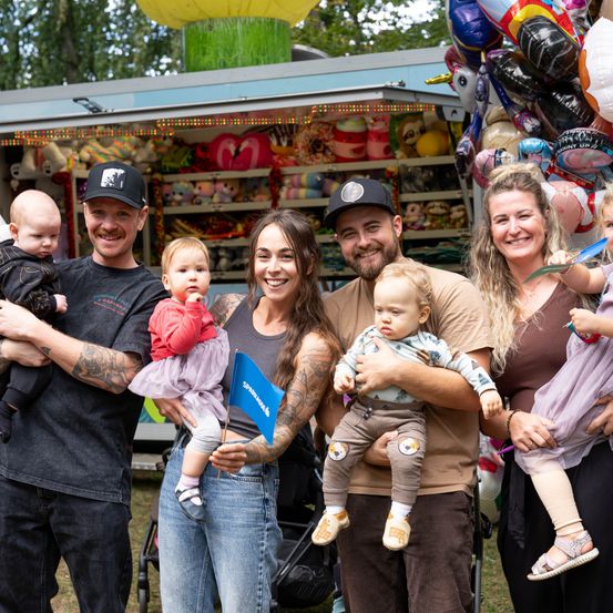 Eine Familie mit fünf Erwachsenen und vier Babys posiert für ein Foto vor einem Jahrmarktstand. Die Erwachsenen lächeln und die Babys werden von ihren Eltern gehalten. Dahinter befinden sich bunte Luftballons und Plüschtiere.