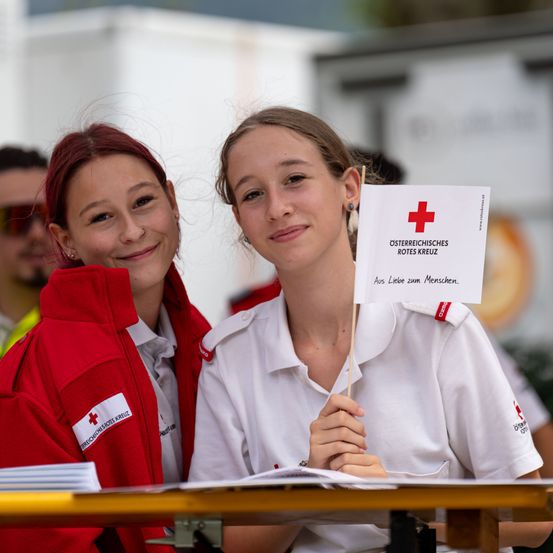 Zwei junge Frauen in Rotkreuz-Uniformen lächeln und posieren für ein Foto. Eine hält ein Schild mit dem Rotkreuz-Logo und einer Botschaft auf Deutsch.