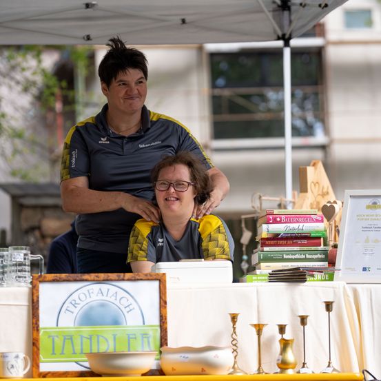 Zwei Frauen stehen hinter einem Tisch auf einem Outdoor-Markt und lächeln. Eine Frau lehnt sich an die andere. Der Tisch hat Bücher, ein Schild und verschiedene Gegenstände.