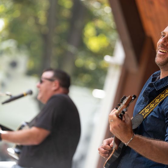 Zwei Männer spielen Gitarren auf einer Bühne mit einem Mikrofon vor ihnen. Der Mann auf der rechten Seite lächelt und schaut in die Kamera.