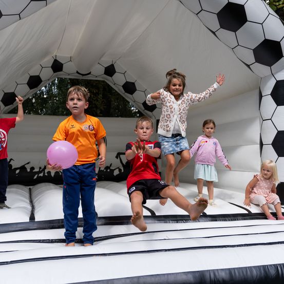 Eine Gruppe von Kindern spielt in einer großen aufblasbaren Fußballstruktur, wobei ein Kind einen rosa Ball hält und andere herumspringen.