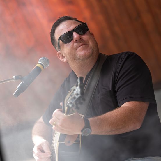 Ein Mann mit Sonnenbrille und schwarzem T-Shirt spielt Gitarre und schaut zur Seite. Er steht vor einem Mikrofon auf einem Ständer.