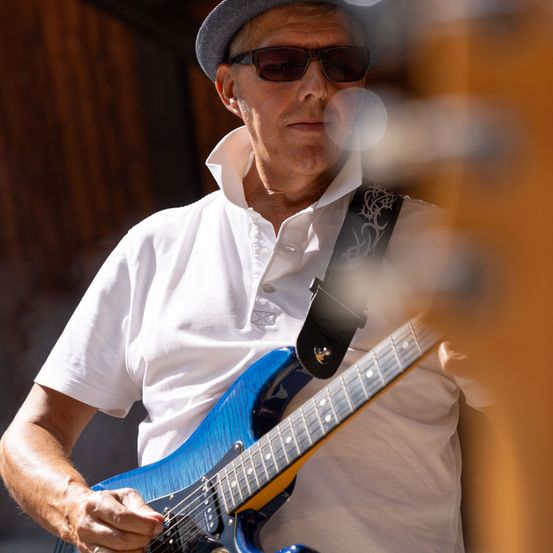 Ein älterer Mann mit Hut und Sonnenbrille spielt eine blaue E-Gitarre. Er trägt ein weißes Poloshirt.