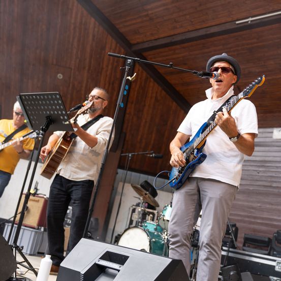 Drei Musiker spielen auf der Bühne, einer spielt eine blaue E-Gitarre, ein anderer eine Gitarre und der dritte Schlagzeug. Sie stehen unter einem Holzdach.