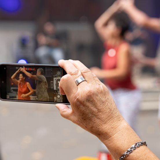 Eine Person filmt eine Tanzaufführung auf ihrem Smartphone, mit einem verschwommenen Hintergrund von Tänzern und einer Bühne.