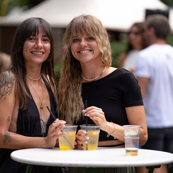 Zwei Frauen lächeln und posieren für ein Foto bei einer Outdoor-Veranstaltung mit Getränken in den Händen.
