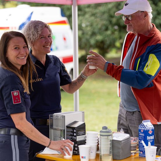 Zwei Frauen in Uniform, eine lächelt und hält einen Becher, stehen hinter einem Tisch mit Erfrischungen. Ein älterer Mann in einem farbenfrohen Jackett lächelt sie an.