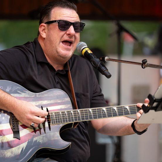 Ein Mann in einem schwarzen Shirt spielt eine Gitarre mit amerikanischem Flaggendesign und singt in ein Mikrofon.
