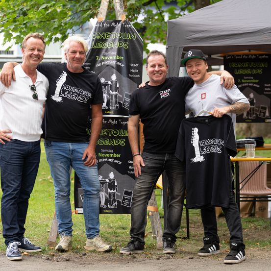 Vier Männer stehen zusammen vor einem Banner, das für die Hometown Legends-Veranstaltung wirbt. Sie alle lächeln und scheinen für ein Foto zu posieren.