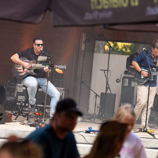Zwei Männer spielen auf der Bühne mit Gitarren, einer sitzt auf einem Hocker, der andere steht, mit einem Publikum im Hintergrund.