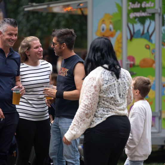 Eine Gruppe von Menschen steht draußen, zwei Männer und zwei Frauen, einer hält ein Getränk. Ein kleiner Junge steht in der Nähe. Dahinter steht ein farbenfroher Imbisswagen.
