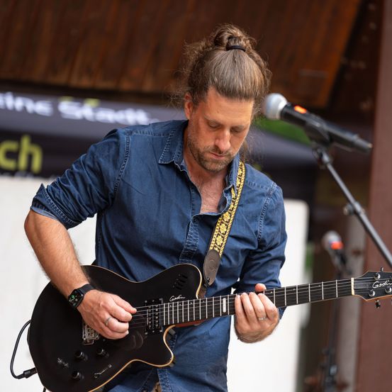 Ein Mann mit Bart und Pferdeschwanz spielt eine schwarze E-Gitarre vor einem Mikrofon. Er trägt ein blaues Hemd und einen gelben Gurt.