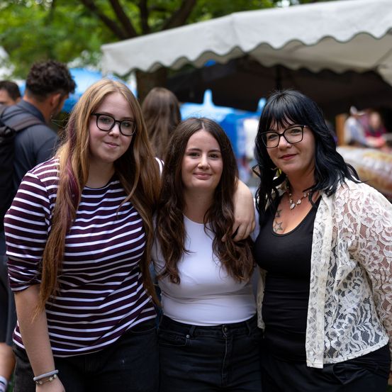 Drei Frauen stehen bei einer Outdoor-Veranstaltung zusammen und lächeln für die Kamera. Die Frau links trägt eine Brille und ein gestreiftes Shirt. Die Frau in der Mitte hat lange Haare und ein weißes Oberteil. Die Frau rechts trägt eine Spitzenjacke und hat ein Tattoo auf ihrer Schulter.