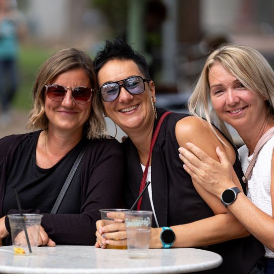 Drei Frauen sitzen zusammen an einem Tisch, lächeln und posieren für ein Foto. Sie tragen Sonnenbrillen und halten Getränke.