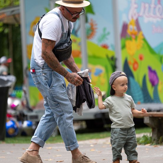 Ein Mann mit Tattoos und Hut hält die Hand eines Babys, während sie in einem Park spazieren gehen. Das Baby trägt einen Hut und schaut etwas an. Ein farbenfrohes Wandbild ist im Hintergrund.