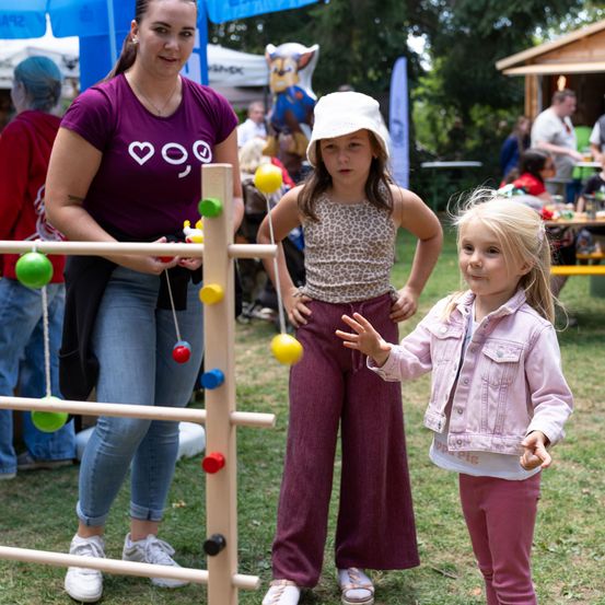 Eine Frau und zwei Mädchen spielen ein Spiel mit bunten Bällen auf einer Holzstruktur in einem Park.