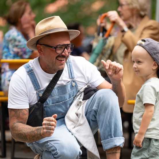 Ein Mann in einer Jeans-Latzhose und einem Hut sitzt auf dem Boden, lächelt und interagiert mit einem kleinen Jungen, der eine Kappe trägt. Dahinter sitzen eine Frau mit einer Handtasche und eine weitere Person.