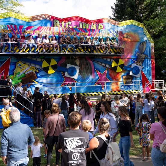 Eine bunte Achterbahn im Freizeitpark mit Menschen, die darauf sitzen, und anderen, die in der Schlange warten. Die Achterbahn ist farbenfroh mit Comic-Charakteren und einem radioaktiven Symbol dekoriert.