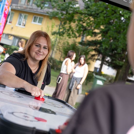 Eine Frau spielt Air Hockey im Freien, mit einem gelben Gebäude und Bäumen im Hintergrund.