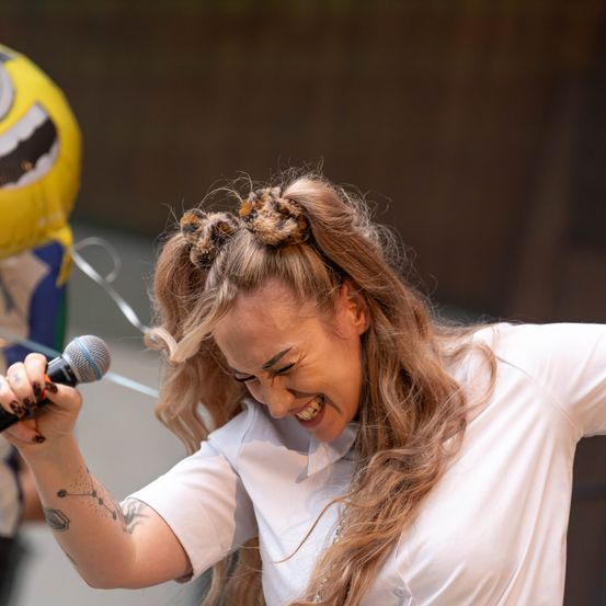 Eine Frau mit Tätowierungen an den Armen singt in ein Mikrofon, während ein Ballon hinter ihr schwebt.