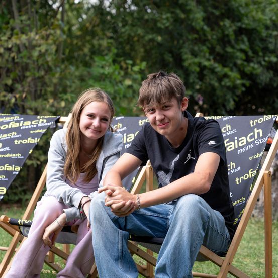 Ein Junge und ein Mädchen sitzen auf Holzstühlen in einem Park, lächeln und posieren für ein Foto. Die Stühle haben Banner mit dem Text 'Trofaiach meine Stadt'. Dahinter sind Bäume.