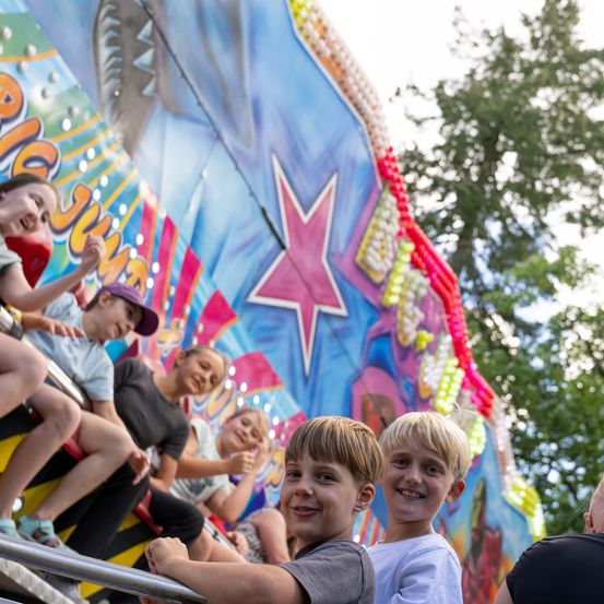 Mehrere Kinder fahren auf einer bunten Karussellfahrt, wobei zwei Jungen im Vordergrund in die Kamera lächeln.