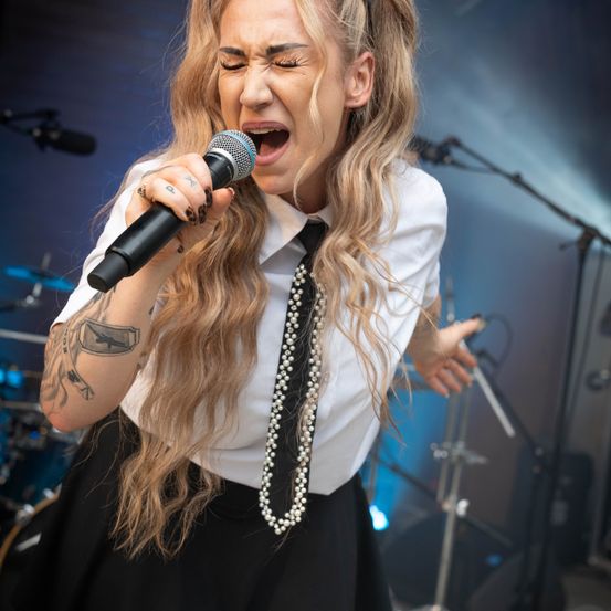 Eine Frau mit langen blonden Haaren singt in ein Mikrofon auf der Bühne, trägt ein weißes Hemd und einen schwarzen Rock.