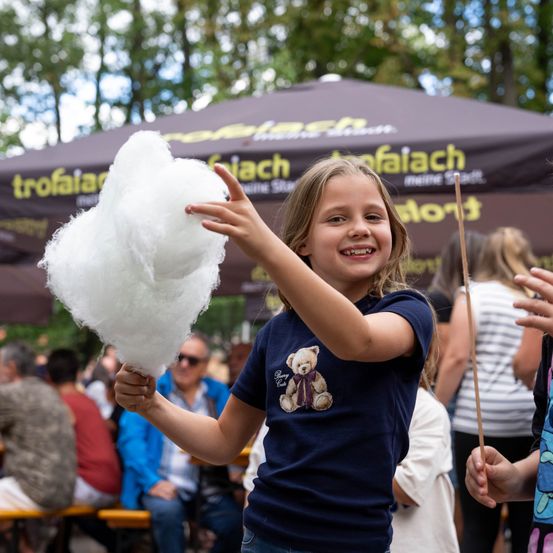 Ein junges Mädchen hält eine große Zuckerwatte in den Händen und lächelt fröhlich bei einer Veranstaltung im Freien. Hinter ihr sitzen Menschen unter einem Zelt, und Bäume sind im Hintergrund zu sehen.