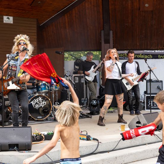 Eine Rockband tritt auf einer Bühne auf, während ein junger Junge davor tanzt. Die Sängerin hält ein Mikrofon, während der Gitarrist eine E-Gitarre spielt. Ein weiterer Gitarrist und ein Schlagzeuger sind im Hintergrund.