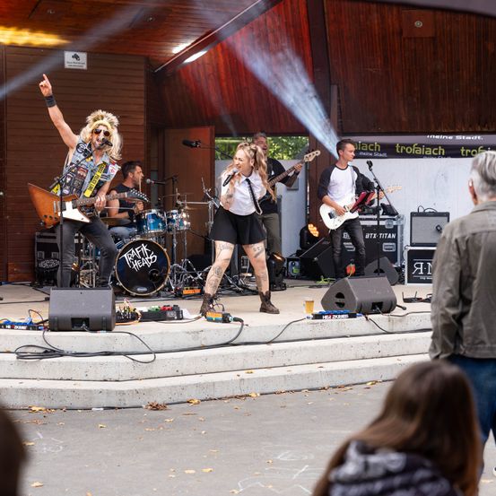 Eine Rockband tritt auf einer Bühne auf, während eine Menge zuschaut. Der Leadsänger steht in der Mitte, mit Gitarristen auf beiden Seiten. Dahinter sind ein Schlagzeug und Lautsprecher zu sehen.