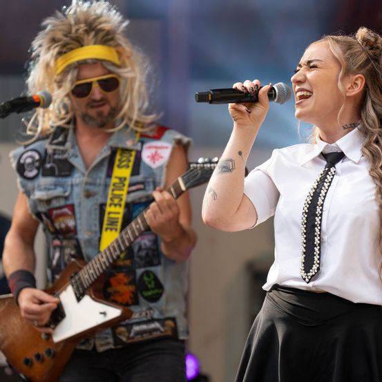 Eine Frau in einem weißen Hemd und einem schwarzen Rock singt in ein Mikrofon, während ein Mann mit einer Gitarre und einem Stirnband hinter ihr spielt.