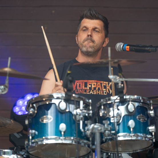 Ein Mann spielt auf der Bühne Schlagzeug und trägt ein schwarzes T-Shirt mit den Worten 'WOLFPACK UNLEASHED'. Er hält Schlagzeugstöcke und sieht konzentriert aus.