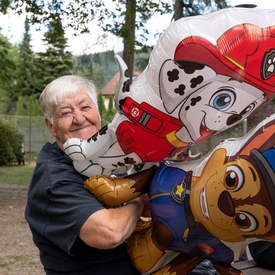 Eine ältere Frau hält große Paw Patrol-Zeichenballons hoch und lächelt in die Kamera.