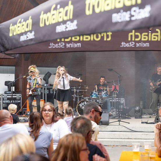 Eine Band tritt auf der Bühne auf, vor einem großen Publikum. Die Sängerin steht in der Mitte, der Gitarrist links. Die Bühne ist mit einem Banner bedeckt.