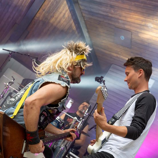 Zwei Männer auf der Bühne spielen Gitarren, einer in einer Jeansweste und der andere in einem weißen Langarmshirt.