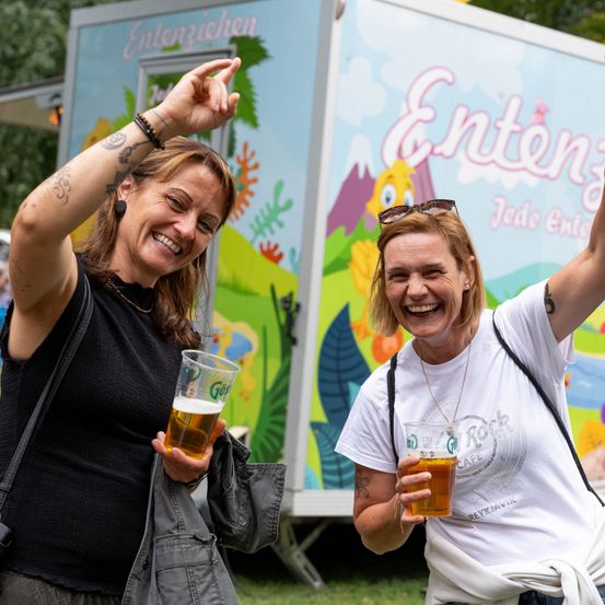 Zwei Frauen, eine mit einer Tätowierung auf dem Arm, lächeln und heben ihre Hände, während sie Bierbecher vor einem Imbisswagen halten.