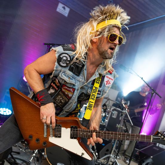 Ein Mann mit langen blonden Haaren, gelbem Stirnband und Sonnenbrille spielt eine E-Gitarre auf der Bühne.