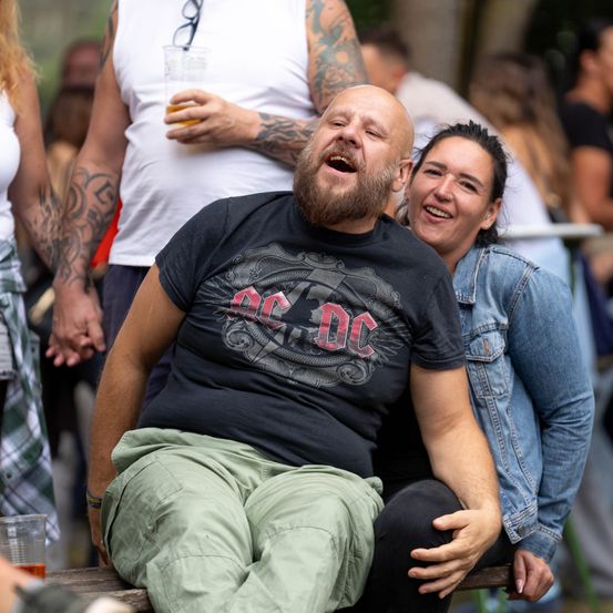 Ein Mann in einem schwarzen AC/DC-T-Shirt lacht und sitzt auf einer Bank mit einer Frau. Sie sind von einer Menge bei einer Veranstaltung im Freien umgeben.