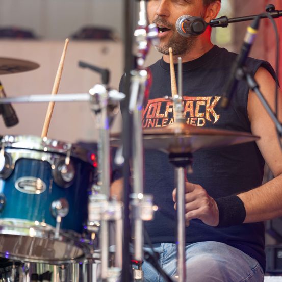 Ein Mann spielt die Drums mit einem Mikrofon vor ihm. Er trägt ein ärmelloses Shirt mit Bandlogo.