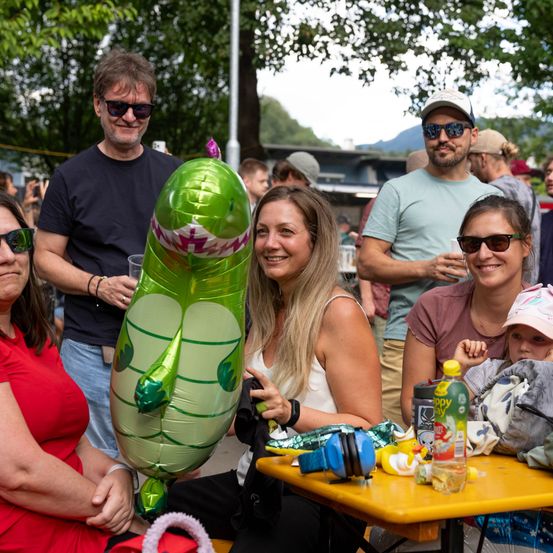 Eine Gruppe von Menschen bei einer Outdoor-Veranstaltung, eine Frau hält einen grünen Ballon in Form eines Dinosauriers.