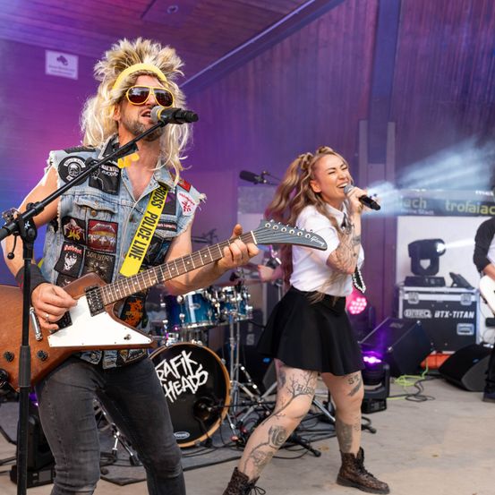 Eine Band tritt auf der Bühne auf, mit einem Gitarristen und einer Sängerin. Der Gitarrist trägt eine Jeansweste mit Aufnähern und eine gelbe Kopfband. Die Sängerin, eine Frau, hält ein Mikrofon und lächelt. Hinter ihnen sind ein Schlagzeug und Lautsprecher zu sehen.