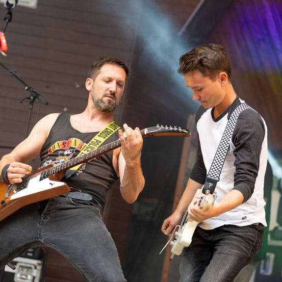 Zwei Männer spielen Gitarren auf der Bühne, einer in einem schwarzen Tanktop und der andere in einem weißen und schwarzen Langarmshirt.