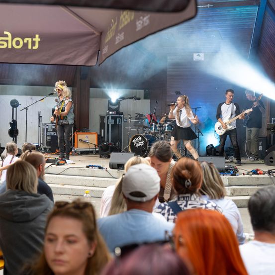 Eine Band tritt auf der Bühne auf, vor einem großen Publikum. Die Sängerin singt in ein Mikrofon, während die Gitarristen ihre Instrumente spielen. Die Bühne ist gut beleuchtet mit Scheinwerfern und Raucheffekten.