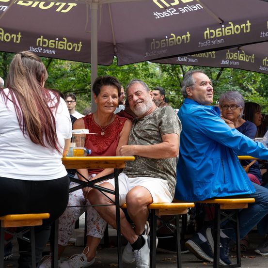 Eine Gruppe von Menschen sitzt unter einem Regenschirm in einem Außenrestaurant. Ein Mann in einem Camouflage-Shirt und eine Frau in einem roten Oberteil lächeln und schauen in die Kamera.