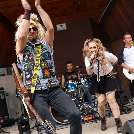 Eine Rockband tritt auf der Bühne auf, ein Mann in einer Jeansweste mit Aufnähern und eine Frau singt in ein Mikrofon.
