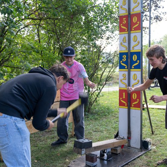 Drei junge Männer nehmen an einem Outdoor-Spiel teil, wobei einer einen Hammer hält und auf ein Ziel schlägt. Das Ziel hat Zahlen und Buchstaben darauf, und die anderen beiden beobachten. Der Hintergrund zeigt Bäume und einen Rasenbereich.