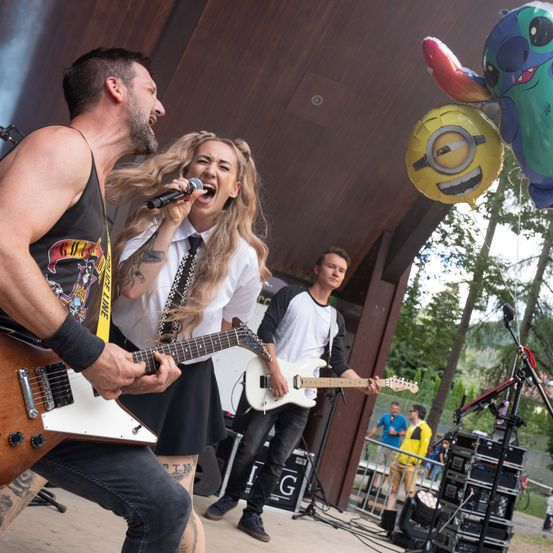 Eine Band tritt auf der Bühne auf, eine Frau singt in ein Mikrofon, ein Mann spielt Gitarre und ein anderer Mann spielt Gitarre. Dahinter befinden sich Luftballons und eine Menge.