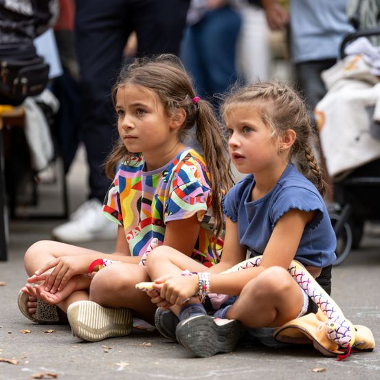 Zwei kleine Mädchen sitzen auf dem Boden, eines trägt ein farbenfrohes Shirt und das andere blau, mit einem Kinderwagen und Menschen im Hintergrund.