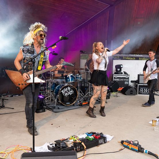 Eine Rockband tritt auf der Bühne auf, mit einem Gitarristen, einem Sänger und einem Schlagzeuger, mit Rauch und Lichtern im Hintergrund.